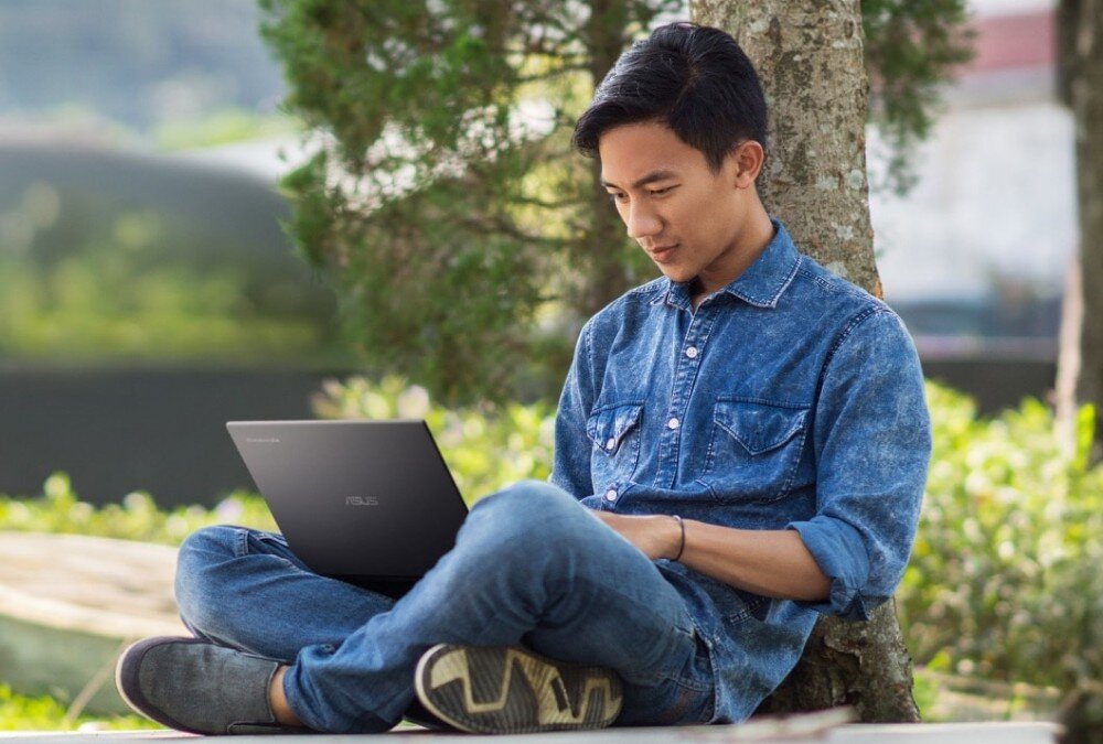 Laptop ASUS Chromebook Plus CX3402CVA Mężczyzna w dżinsowej koszuli siedzi pod drzewem na trawie, trzymając otwarty ciemny laptop na kolanach. Patrzy na ekran, a w tle widoczne są drzewa i rozmyte elementy krajobrazu, procesor, pamięć, dysk, wydajność