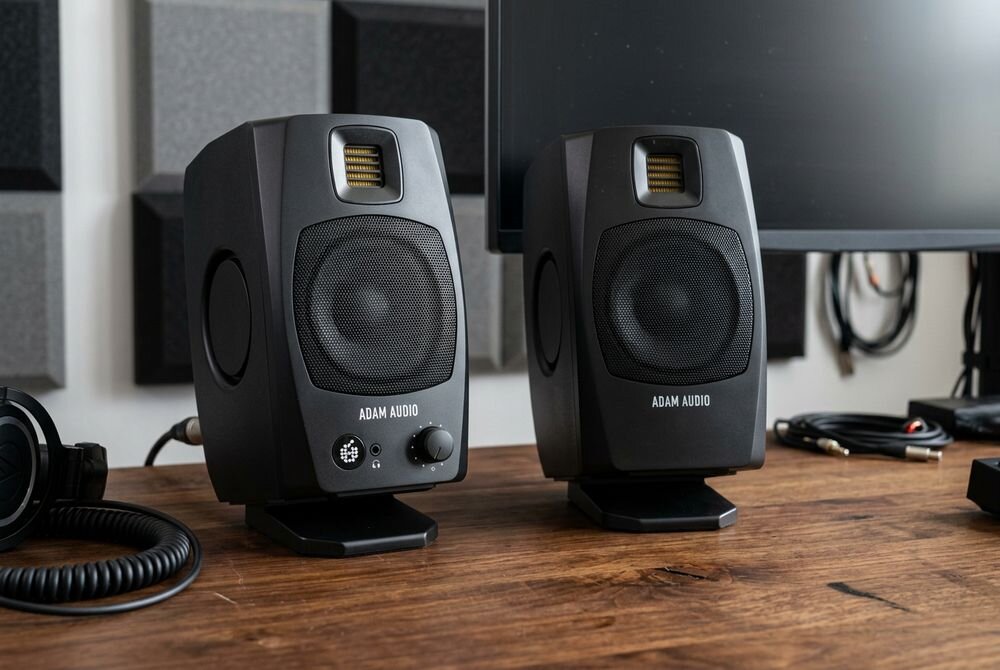 Na drewnianym biurku stoją dwa czarne monitory aktywne z logo ADAM AUDIO. W tle widoczny jest fragment monitora komputerowego oraz kable i słuchawki.