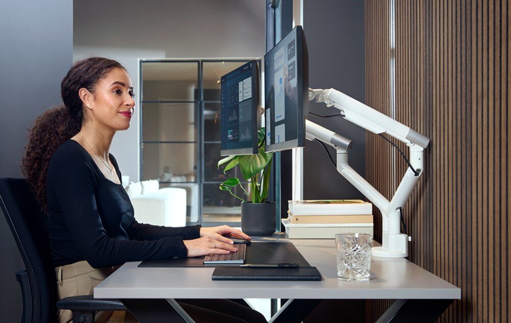 Uchwyt do monitora ONE FOR ALL DM7220 Kobieta siedząca przy biurku i pracująca na komputerze z dwoma monitorami zamontowanymi na podwójnym uchwycie biurkowym