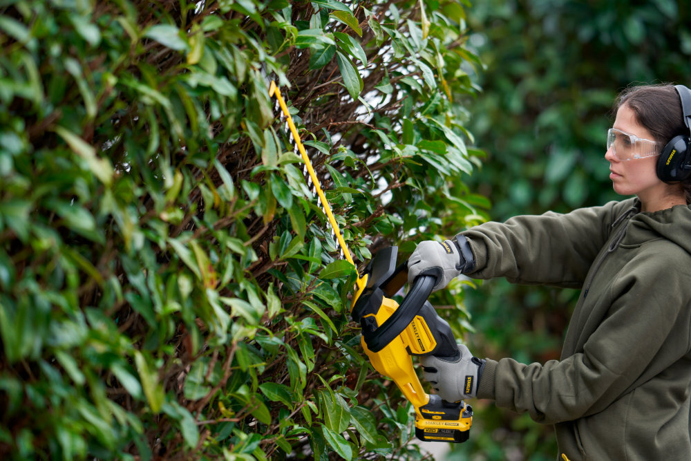Nożyce do żywopłotu STANLEY SFMCHT855M1-QW 550mm 18V akumulatorowe ogród osoba przycina żywopłot osoba widoczna bokiem w tle inne rośliny silnik szczotkowy stabilna moc podczas pracy nad żywopłotem narzędzie łatwe w utrzymaniu i przewidywalne w działaniu bez nagłych spadków prędkości cięcia równo i płynnie komfort i efektywność