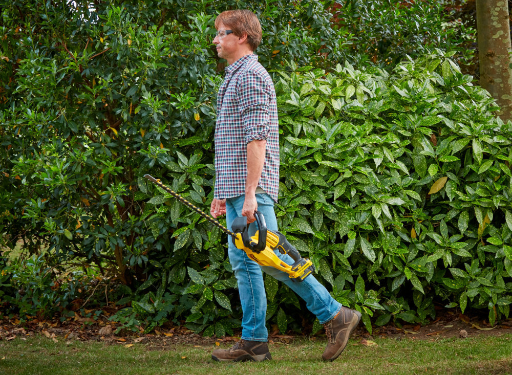 Nożyce do żywopłotu STANLEY SFMCHT855M1-QW 550mm 18V akumulatorowe ogród osoba idzie po trawniku z nożycami w dłoni w tle rośliny waga około 2,9 kg zmniejsza obciążenie ramion użytkownik odczuwa większy komfort