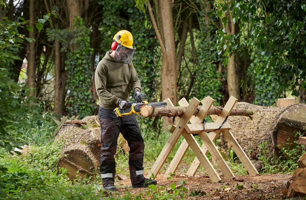 Piła akumulatorowa STANLEY 18V Fatmax V20 SFMCCS730B-XJ las osoba tnie pień drzewa w tle drzewa liście osoba w żółtym kasku wokół powalone drzewa zębatki oporowe zwiększają przyczepność pilarki do ciętego materiału waga około 4,3 kg z akumulatorem 4,0 Ah stabilność swoboda manewrowania
