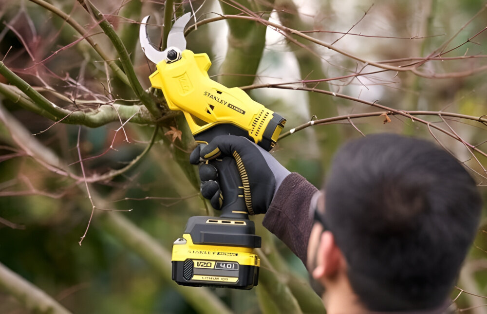 Sekator do gałęzi akumulatorowy STANLEY FatMax SFMCPP32M1-QW ogród osoba przycina gałęzie na krzewie widok od tyłu do pielęgnacji roślin przycinanie mokrego i suchego drewna ogranicza obciążenie dłoni i nadgarstków ułatwia regularne prace porządkowe poprawia komfort codziennej pielęgnacji zieleni