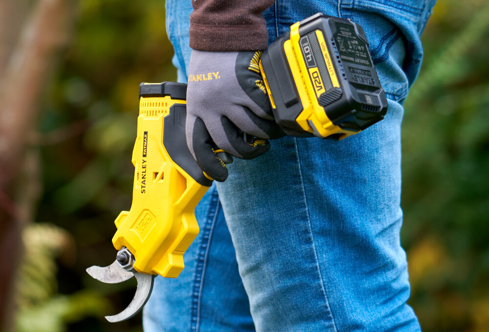 Sekator do gałęzi akumulatorowy STANLEY FatMax SFMCPP32M1-QW ogród osoba trzyma sekator w dłoni tuż przy nodze rozmazane tło napęd elektryczny przejmuje siłę potrzebną do cięcia zmniejsza obciążenie dłoni, stawów i przedramion sprzyja dłuższej pracy ciągłej lepszą kontrolę nad narzędziem do 500 cięć na jednym ładowaniu