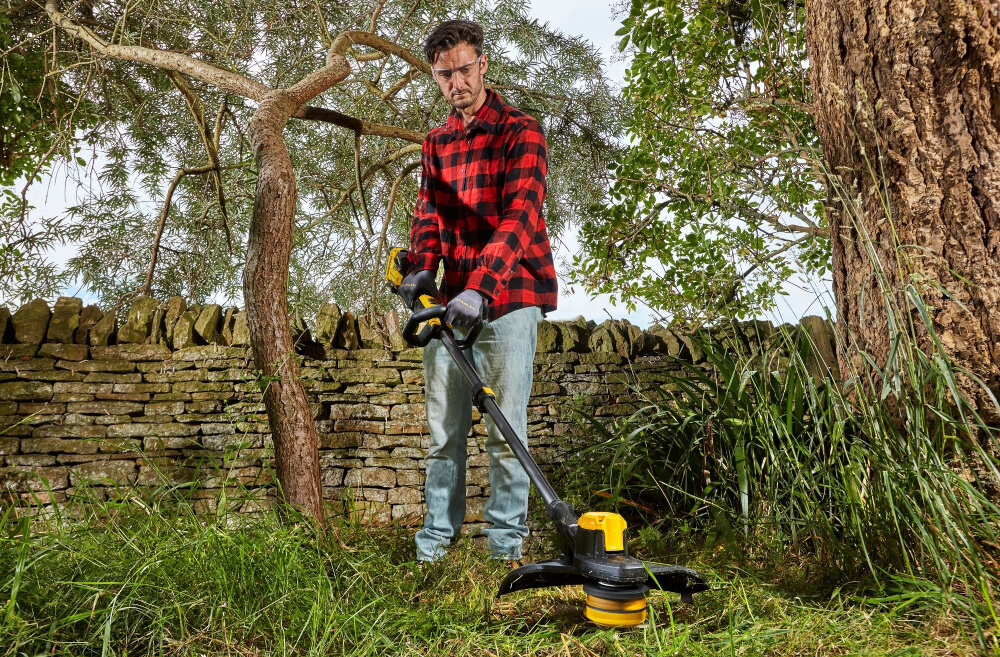 Podkaszarka akumulatorowa STANLEY SFMCSTB933B-XJ ogród osoba kosi trawę w tle drzewa murek niebo zadbany ogród pielęgnacja ogrodu do pracy w trudno dostępnych miejscach