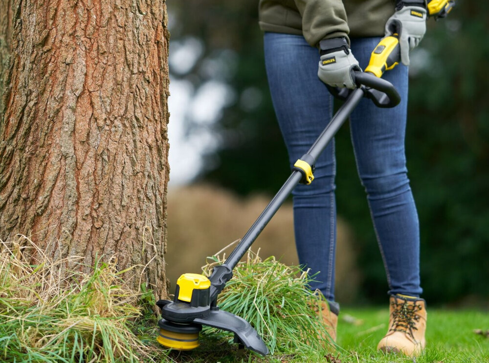 Podkaszarka akumulatorowa STANLEY SFMCSTB933B-XJ ogród osoba podkasza trawę wokół drzewa rozmazane tło przekładnia zębata zwiększa moment obrotowy skuteczniejsze cięcie równa linia koszenia mniejsze ryzyko zapychania się elementu tnącego
