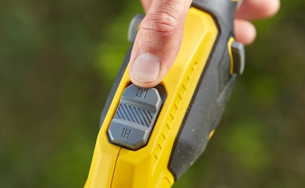 Podkaszarka akumulatorowa STANLEY SFMCSTB933B-XJ zbliżenie na przycisk do regulacji prędkości rozmazane tło regulacja wysokiej i niskiej prędkości zmienna prędkość obrotowa do 6000 obr/min można dopasować tempo pracy do rodzaju roślinności