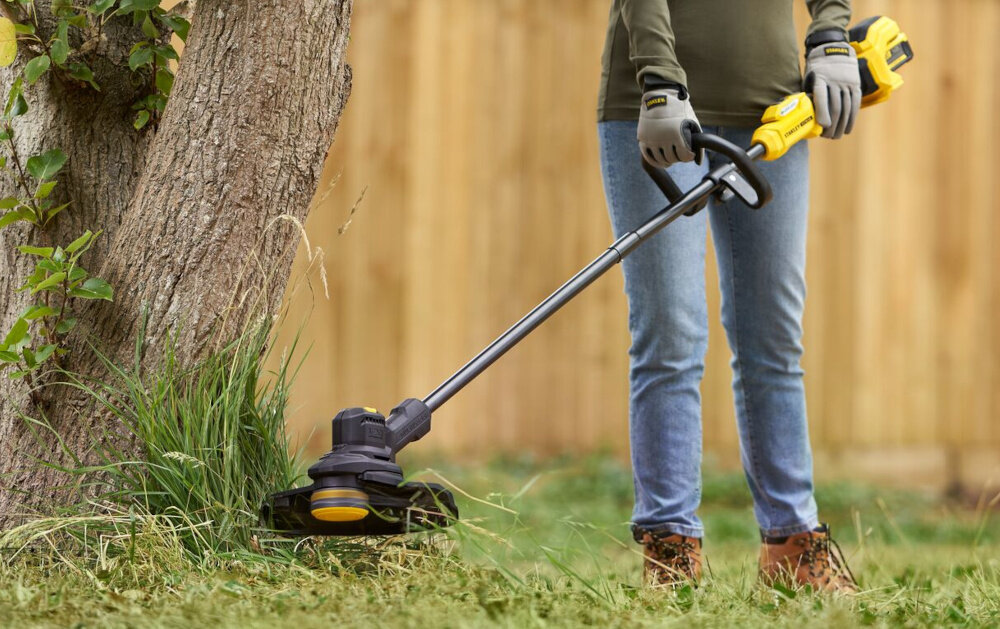 Podkaszarka akumulatorowa STANLEY SFMSTB930B-XJ ogród osoba kosi trawę w tle drzewa drewniany płot niebo zadbany ogród pielęgnacja ogrodu do pracy w trudno dostępnych miejscach