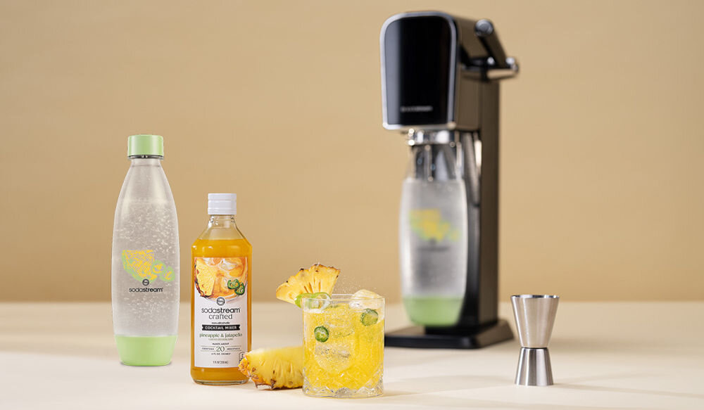 Zestaw upominkowy SODASTREAM Crafted Ananas & Jalapeño jasne tło z tyłu stoi saturator butelka zielony spód zielona nakrętka żółty napój w szklance kawałki ananasa papryczki kostki lodu wielorazowa butelka mniej plastiku tritan do wielokrotnego użytkowania bezpieczeństwo wolna od BPA możliwość mycia w zmywarce świeżość na dłużej szczelny korek do 4 lat użytkowania kompatybilność modele Terra, Art, Duo, Enso, Spirit i Source