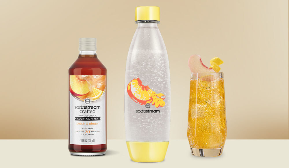 Zestaw upominkowy SODASTREAM Crafted Brzoskwinia & Imbir jasne tło butelka żółty spód żółta zakrętka rysunek na butelce obok stoi syrop w szklanej butelce oraz żółty napój bąbelkowa przyjemność
