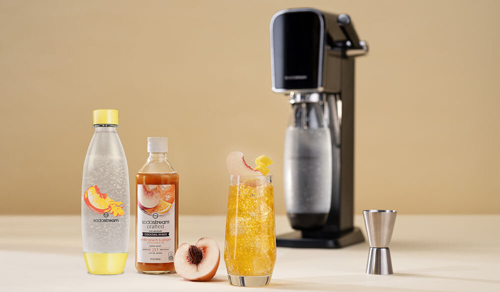 Zestaw upominkowy SODASTREAM Crafted Brzoskwinia & Imbir jasne tło z tyłu stoi saturator butelka żółty spód żółta nakrętka pomarańczowy napój w szklance kawałki brzoskwini kostki lodu wielorazowa butelka mniej plastiku tritan do wielokrotnego użytkowania bezpieczeństwo wolna od BPA możliwość mycia w zmywarce świeżość na dłużej szczelny korek do 4 lat użytkowania kompatybilność modele Terra, Art, Duo, Enso, Spirit i Source