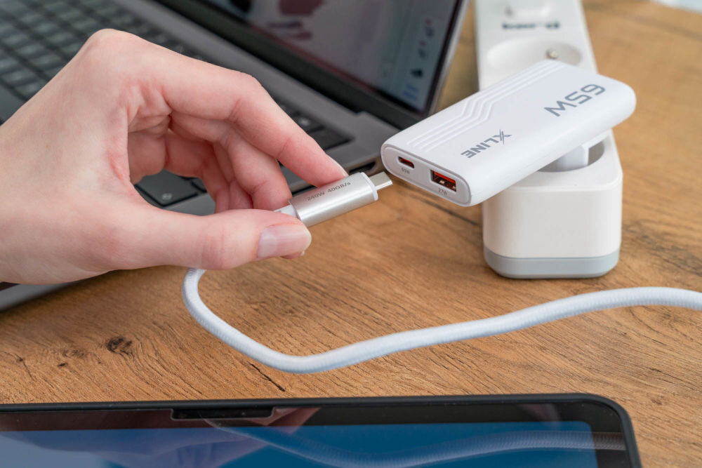 Osoba trzyma kabel USB-C, który jest podłączany do białej ładowarki sieciowej z napisem \'XLINE 65W\'. Ładowarka jest wpięta do listwy zasilającej, a w tle widać laptopa.