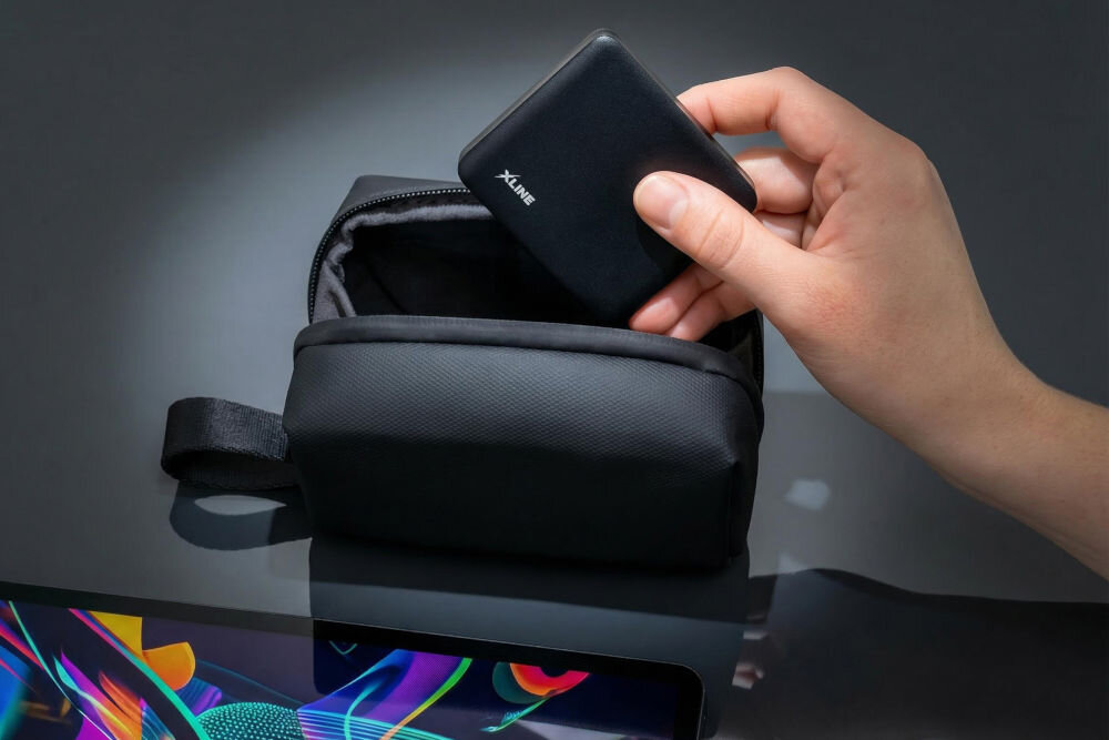 Powerbank XLINE XPB305K 5000 mAh 20W Osoba wkładająca bardzo mały powerbank X-LINE do czarnej torby podróżnej, podkreślająca kieszonkowy rozmiar urządzenia typu 