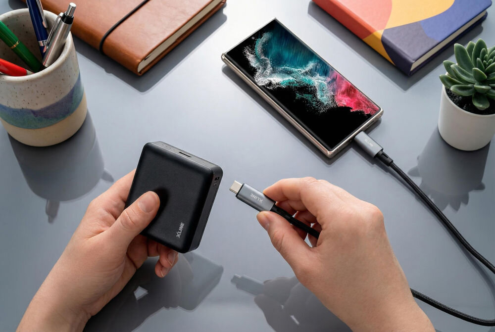 Powerbank XLINE XPB310K 10000 mAh 22.5W Dłoń podłączająca kabel USB-C do powerbanku X-LINE XPB310K; obok ładujący się smartfon z nowoczesnym wyświetlaczem, widok z góry.