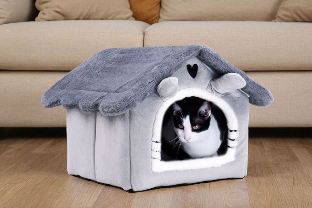 Legowisko dla kota ZWIERZOPASJA Jasper z antypoślizgową podstawą zapobiegającą przesuwaniu Pluszowy, szary domek-legowisko dla kota na drewnianej podłodze przed beżową kanapą. W wejściu do domku siedzi kot czarno-biały.
