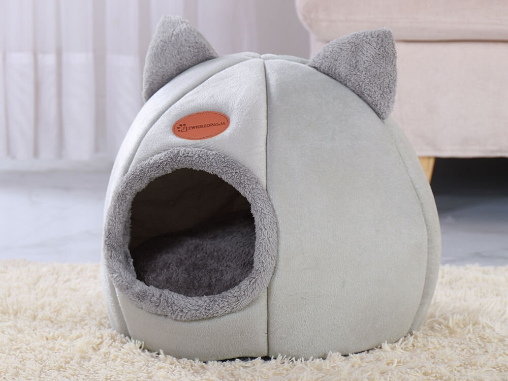 Legowisko dla kota ZWIERZOPASJA Amber szary miękka tkanina wygoda ocieplenie małe pieski koty wyjmowana poduszka 37x37x33 cm Szary, pluszowy domek dla kota w kształcie kopuły z zabawnymi uszami na szczycie. Ma okrągłe wejście obszyte miękkim, futrzanym materiałem i brązową naszywkę z logo ZWIERZOPASJA. Domek stoi na jasnym, puszystym dywanie.