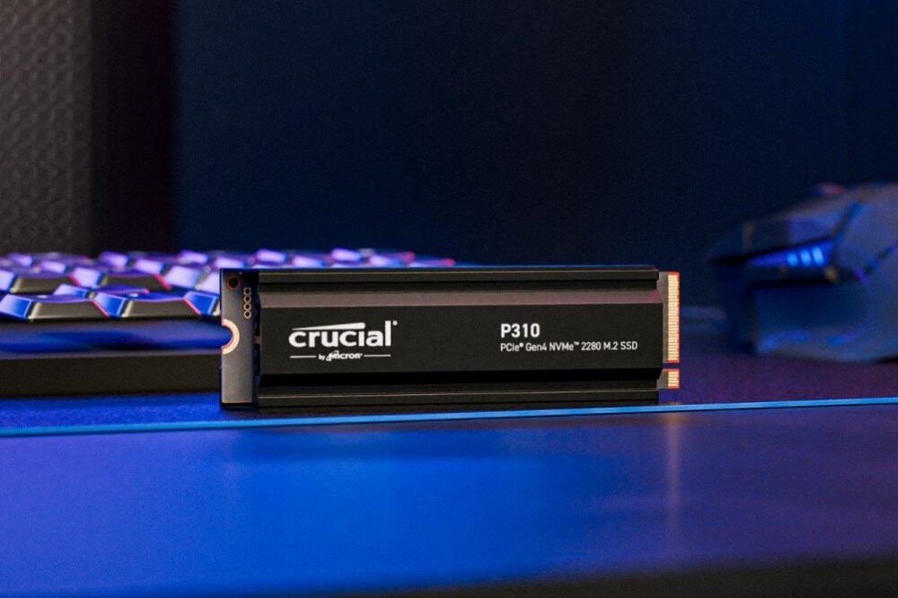 Dysk CRUCIAL P310 Dysk SSD z napisem 'crucial P310 PCIe Gen4 NVMe M.2 SSD' leży na niebieskim biurku, w tle klawiatura i myszka, opis