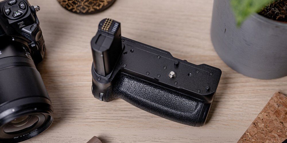 Rzut z góry na Battery Pack Newell MB-N12 do Nikona na drewnianym blacie w otoczeniu aparatu fotograficznego i doniczki. kv początek opisu