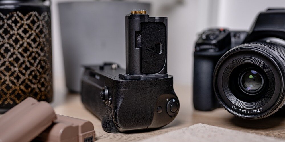 Czarny grip marki Newell MB-N12 do Nikona na drewnianym blacie w otoczeniu lustrzanki i akumulatorów.