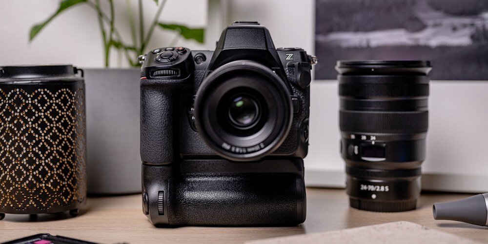 Grip marki Newell MB-N12 przymocowany do aparatu Nikon Z8. Obok obiektyw. W tle roślina doniczkowa.