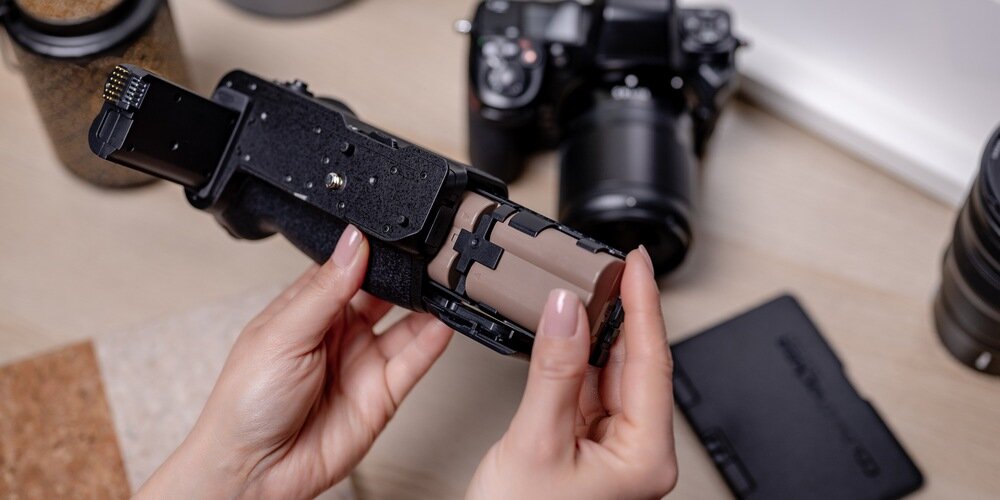 Kobieca dłoń wymienia nad drewnianym blatem akumulator w gripie Newell MB-N12. W tle między innymi aparat fotograficzny