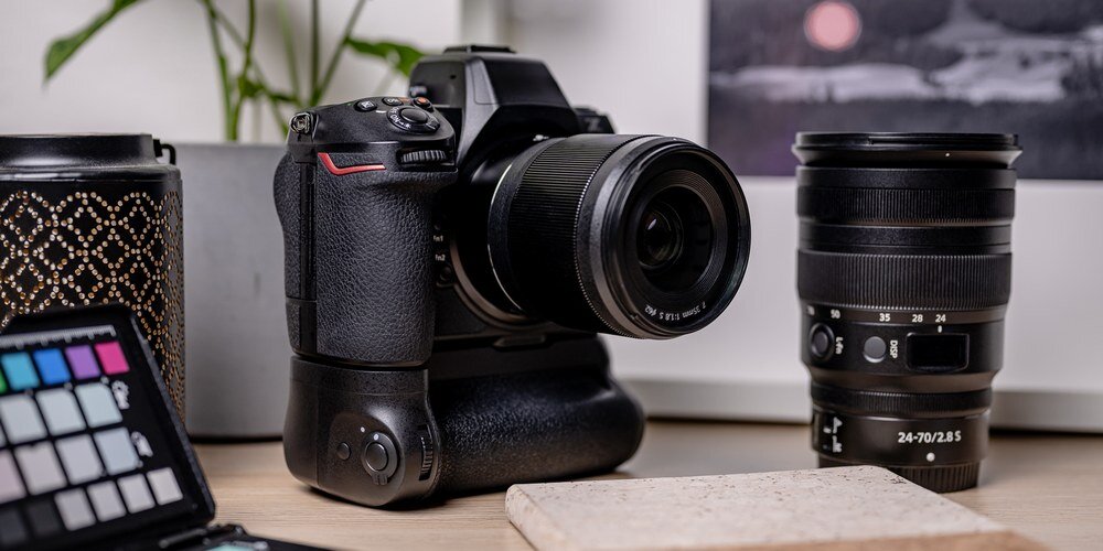 Lustrzanka Nikon Z8 z przymocowanym gripem MB-N12 na drewnianym blacie. Z lewej strony stoi lampion, z prawej obiektyw.
