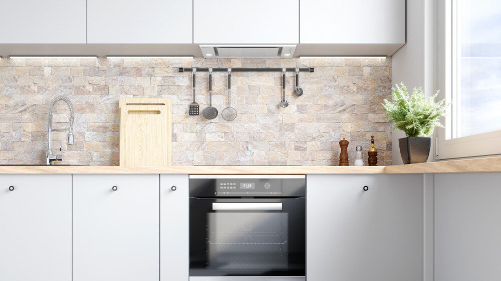OKAP GLOBALO DESOLERO 60.3 Jasna kuchnia z okapem do zabudowy umieszczonym pod górnymi szafkami na tle ściany z kamiennych płytek, z piekarnikiem w zabudowie poniżej, deską do krojenia, relingiem z akcesoriami i doniczką przy oknie