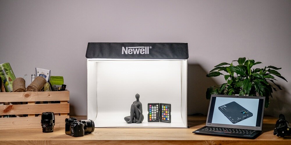 Na drewnianym stole znajduje się namiot bezcieniowy z napisem \'Newell\', wewnątrz którego umieszczono figurkę i wzornik kolorów. Obok namiotu stoi laptop z wyświetlonym obrazem oraz roślina doniczkowa.