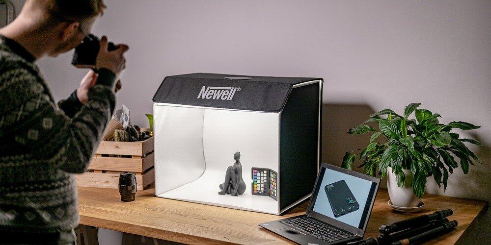 Na drewnianym stole znajduje się namiot bezcieniowy z logo \'Newell\', wewnątrz którego umieszczono figurkę i kartę kolorów. Obok namiotu stoi otwarty laptop, a w tle widoczna jest roślina doniczkowa.