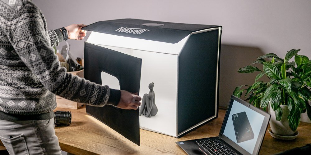 Osoba ustawia czarny panel w namiocie bezcieniowym z widocznym logo \'Newell\'. Obok znajduje się laptop z wyświetlonym zdjęciem oraz roślina doniczkowa.
