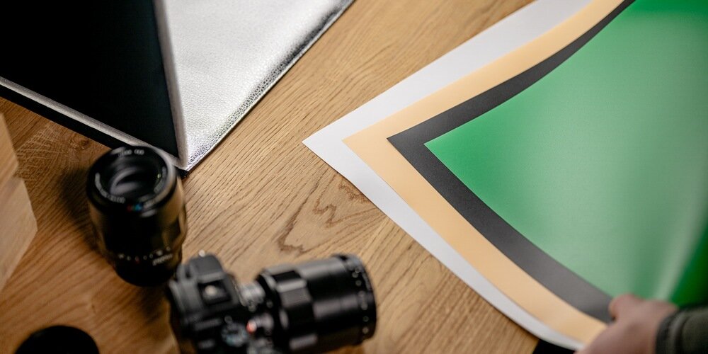 Na drewnianym stole leży aparat fotograficzny, obiektyw oraz kolorowe arkusze papieru w odcieniach zieleni, pomarańczu i czerni. W tle widoczna jest srebrna powierzchnia, prawdopodobnie część namiotu bezcieniowego.
