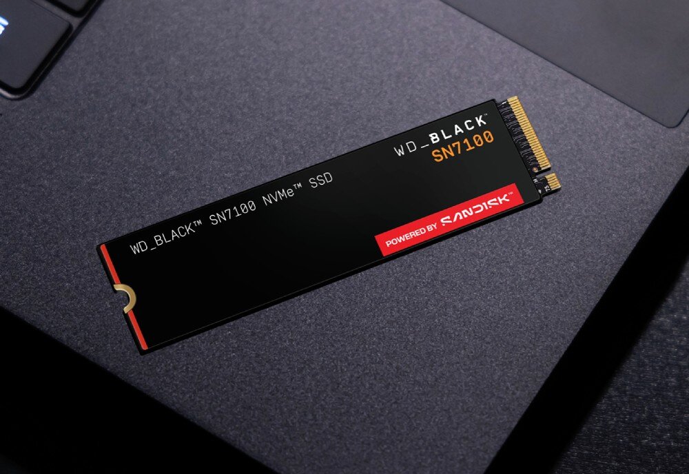 Dysk SANDISK WD Black SN7100 Podłużny dysk SSD M.2 z napisami 'WD_BLACK SN7100 NVMe SSD' oraz logotypem 'powered by SanDisk' leży na matowej powierzchni laptopa, opis