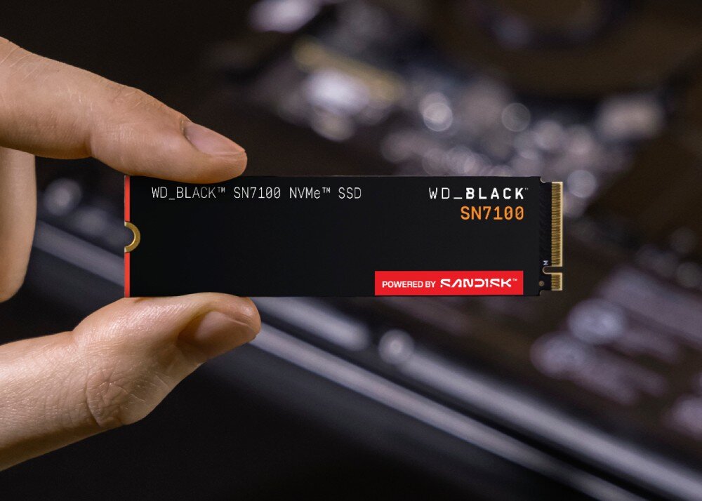 Dysk SANDISK WD Black SN7100 Ludzka dłoń trzyma poziomo dwoma palcami podłużny dysk SSD M.2 z widocznymi napisami i pozłacanymi stykami na tle rozmytej elektroniki, pojemność, format, prędkość, interfejs