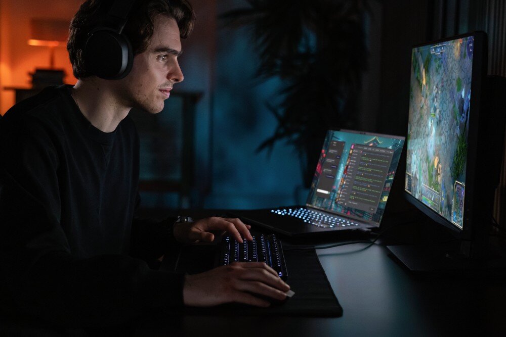 Dysk SANDISK WD Black SN7100 Profil młodego mężczyzny w słuchawkach grającego na komputerze w ciemnym pokoju, przed nim podświetlana klawiatura, mysz oraz dwa monitory, przestrzeń na pliki