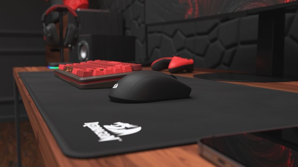 Na drewnianym biurku znajduje się czarna mysz komputerowa na dużej podkładce z logo \'Redragon\'. W tle widoczna jest czerwona klawiatura mechaniczna oraz słuchawki na stojaku.