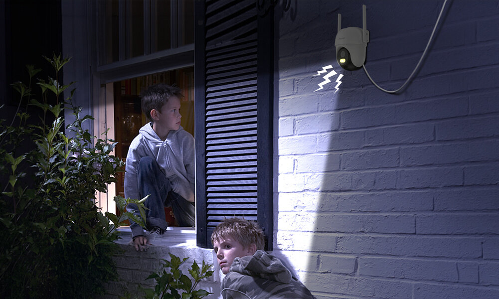 Dwóch zaniepokojonych chłopców wygląda nocą przez otwarte okno. W tle widać zamontowaną na ceglanej ścianie kamerę. Włączony reflektor ich oświetla, a dodatkowo kamera wydaje dźwięk alarmowy.