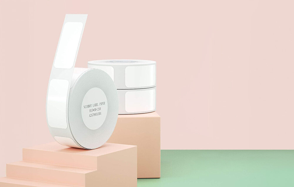 Na obrazie widoczne są trzy rolki białych etykiet termicznych ustawione na pastelowych blokach. Na jednej z rolek widnieje tekst: \'NIIMBOT LABEL PAPER 15*30*210 210PCS/ROLL\'.
