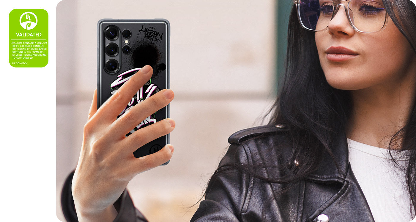Osoba trzyma Galaxy S25 Ultra z założonym etui Rugged Case. Smartfon widziany jest od tyłu, z dłońmi trzymającymi wzory na obudowie. Po lewej stronie widoczny jest znak walidacji ochrony środowiska UL Environmental Claim Validation Mark z napisem 'EF-MS938 ZAWIERA MINIMUM 3% ZAWARTOŚCI BIOLOGICZNEJ, SKŁADAJĄC SIĘ Z 9% ZAWARTOŚCI BIOLOGICZNEJ W RAMACH EF-JS938, TESTOWANE ZGODNIE Z NORMĄ ASTM D6866-22. UL.COM/ECV