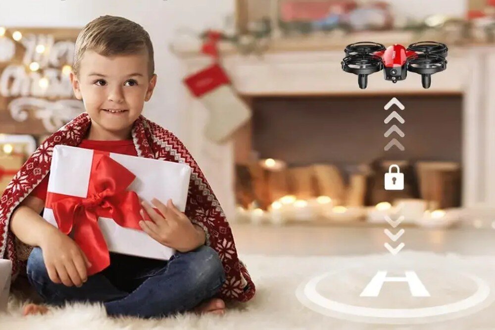 Dron EXTRALINK A24 Chłopiec siedzący na puszystym dywanie trzyma zapakowany prezent, a przed nim w powietrzu unosi się mały czerwono-czarny dron nad grafiką lądowiska z literą 'H'