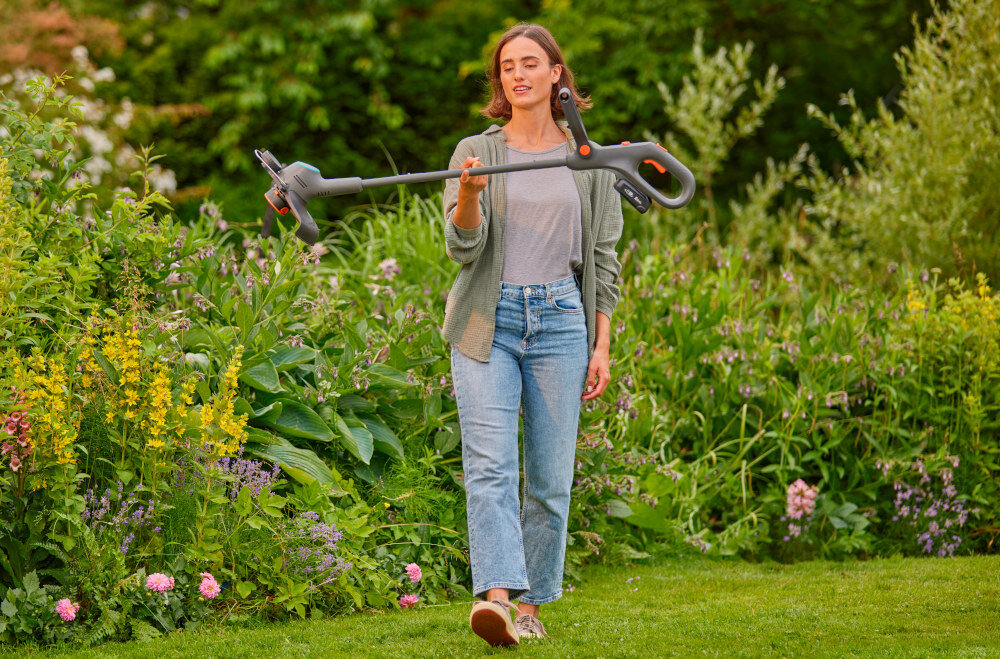 Podkaszarka akumulatorowa GARDENA EasyTrim 14704-20 ogród kwiaty drzewa kobieta niesie podkaszarkę na jednym palcu pielęgnacja trawnika przy krawędziach wokół drzew i rabat szerokość cięcia do 25 cm brak przewodu swoboda ruchów niska masa 2,06 kg z akumulatorem