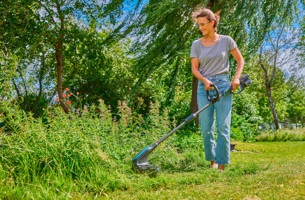 Podkaszarka akumulatorowa GARDENA 14703-20 kobieta tnie wysoką trawę w tle drzewa trawnik funkcja PowerBoost automatycznie zwiększa siłę cięcia w trudniejszych warunkach poprawia tempo pracy