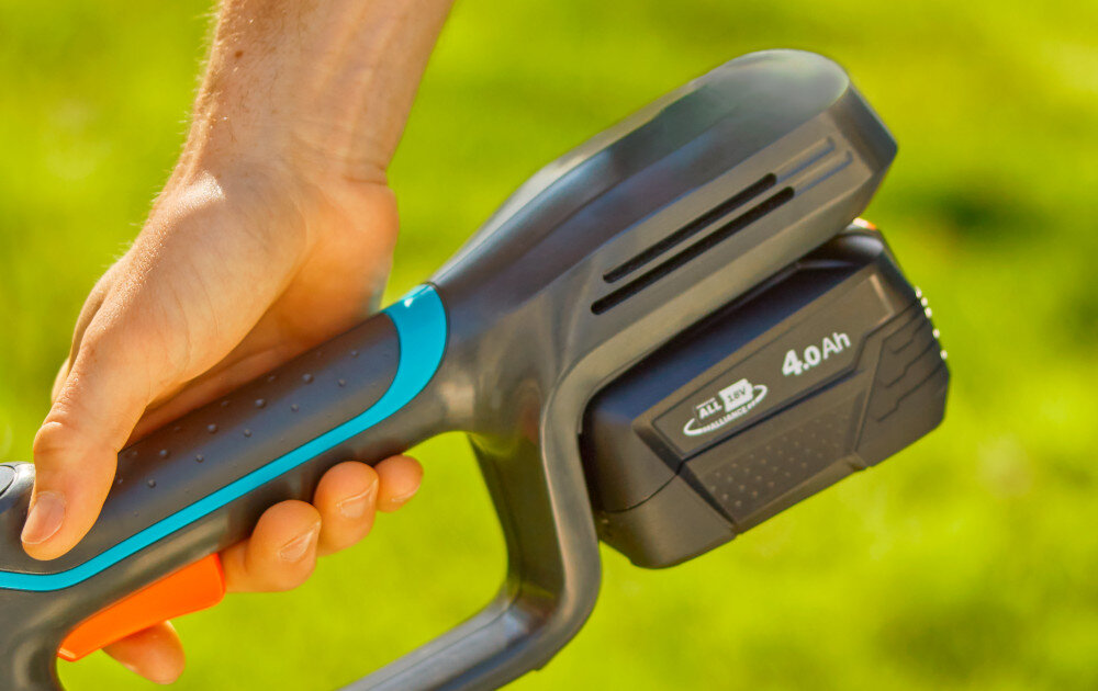 Podkaszarka akumulatorowa GARDENA PowerTrim 30 14703-55 zbliżenie na akumulator osoba trzyma podkaszarkę w dłoniach w tle trawnik zasilana jest akumulatorem 18 V o pojemności 4 Ah system POWER FOR ALL do 40 minut pracy na jednym ładowaniu pasuje również do innych narzędzi ogrodowych i domowych tej samej serii zaoszczędzić na zakupie