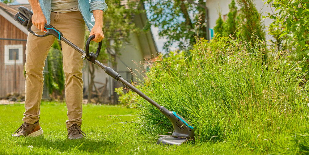 Podkaszarka akumulatorowa GARDENA PowerTrim 30 14703-55 osoba tnie wysoką trawę w tle dom w 50% z plastiku poużytkowego funkcjonalność ekologiczna odpowiedzialność hałas na poziomie 79 dB przy uchu operatora niski poziom wibracji 0,7 m/s² komfort i małe zmęczenie rąk