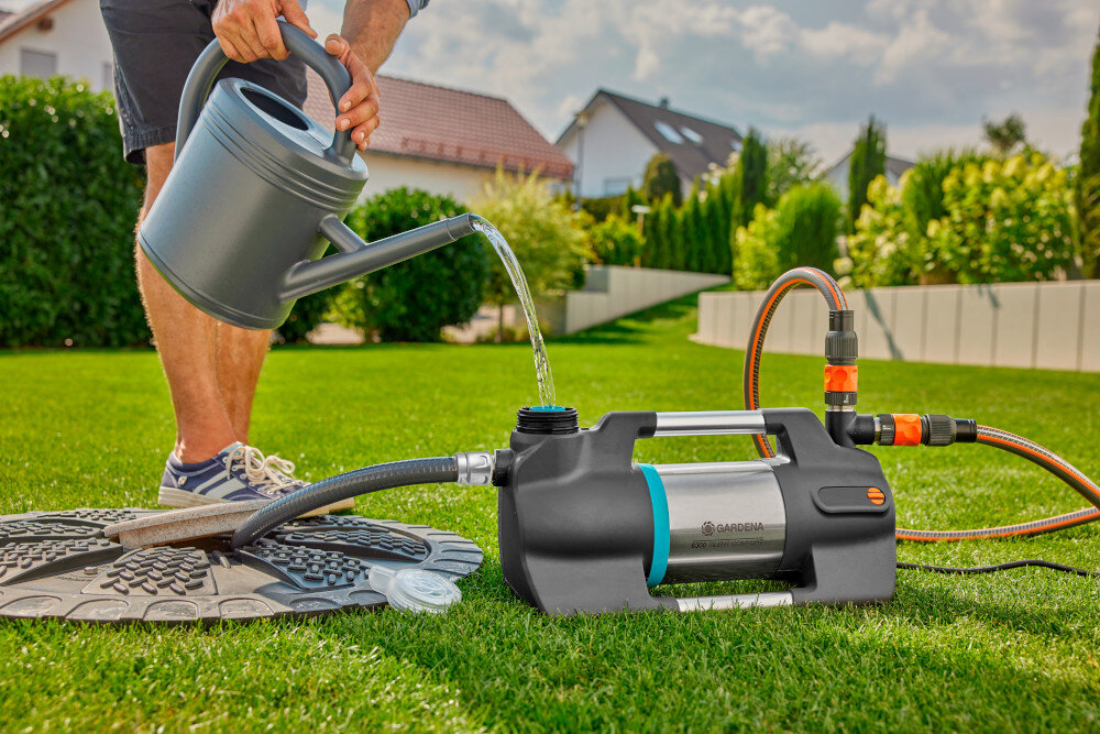 Wąż ssący GARDENA 9092-20 4 m ogród wąż umieszczony w studzience kanalizacyjnej pompuje wodę do pompy osoba dolewa wodę do pompy w tle dom długość 4 metry z materiałów odpornych na podciśnienie mróz i promieniowanie UV cały rok