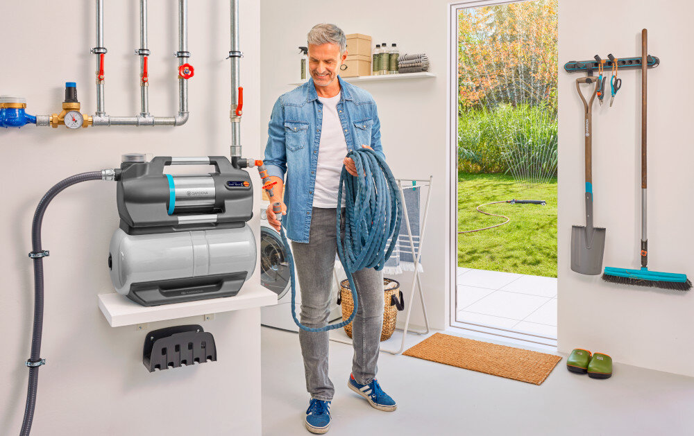 Wąż ssący GARDENA 9092-20 4 m pomieszczenie wąż podłączony do pompy wodnej obok stoi osoba w tle otwarte drzwi widoczny ogród wytrzymała metalowa nakrętka przyłączeniowa bezpieczne i szczelne połączenie eliminacja wycieków i przedostawania się powietrza do systemu komfort użytkowania