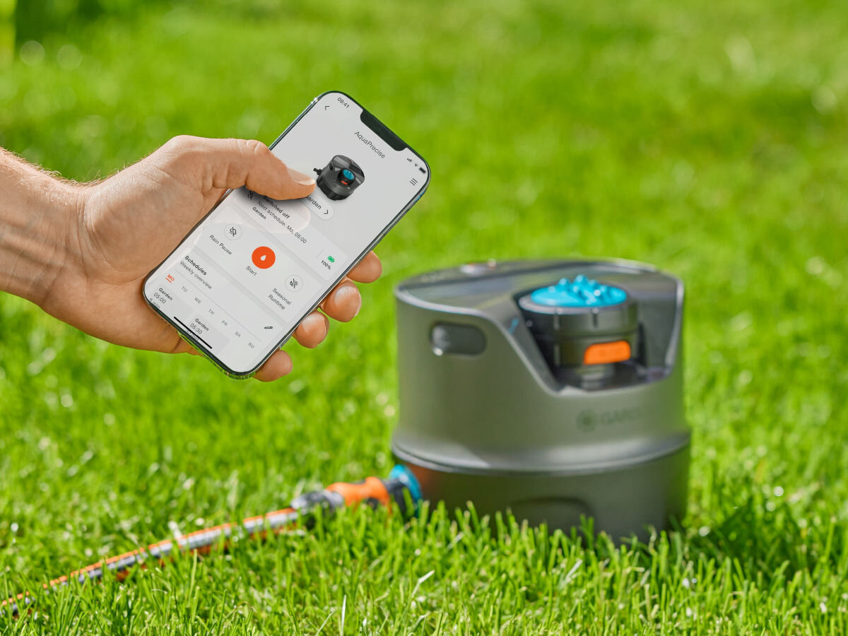 Zraszacz GARDENA AquaPrecise 16000-29, baterie, światło, słoneczne, ładowanie, USB, Aplikacja, dłoń trzyma telefon i aplikację gardena, obok zraszacz zamontowany do trawnika