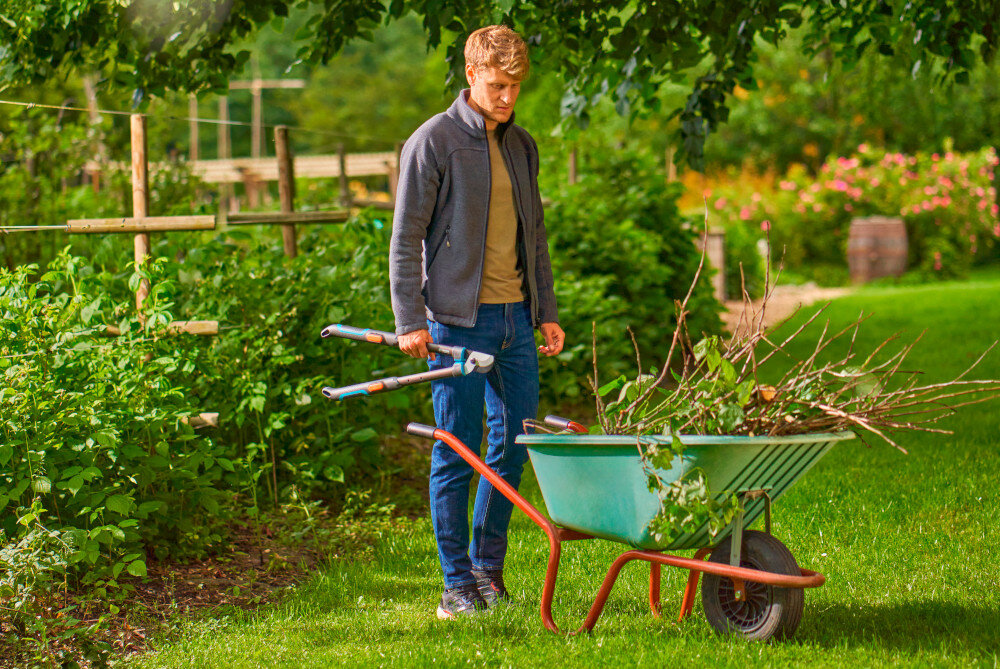 Nożyce do gałęzi GARDENA TeleCut Pro S 12040-20 ogród drzewa osoba stoi na trawniku z nożycami w dłoni obok taczka rabaty kwiaty krzewy długość 520 mm odpowiedni zasięg przy pracy wśród gęstych gałęzi dwuostrzowa konstrukcja ergonomiczny uchwyt lekka aluminiowa budowa
