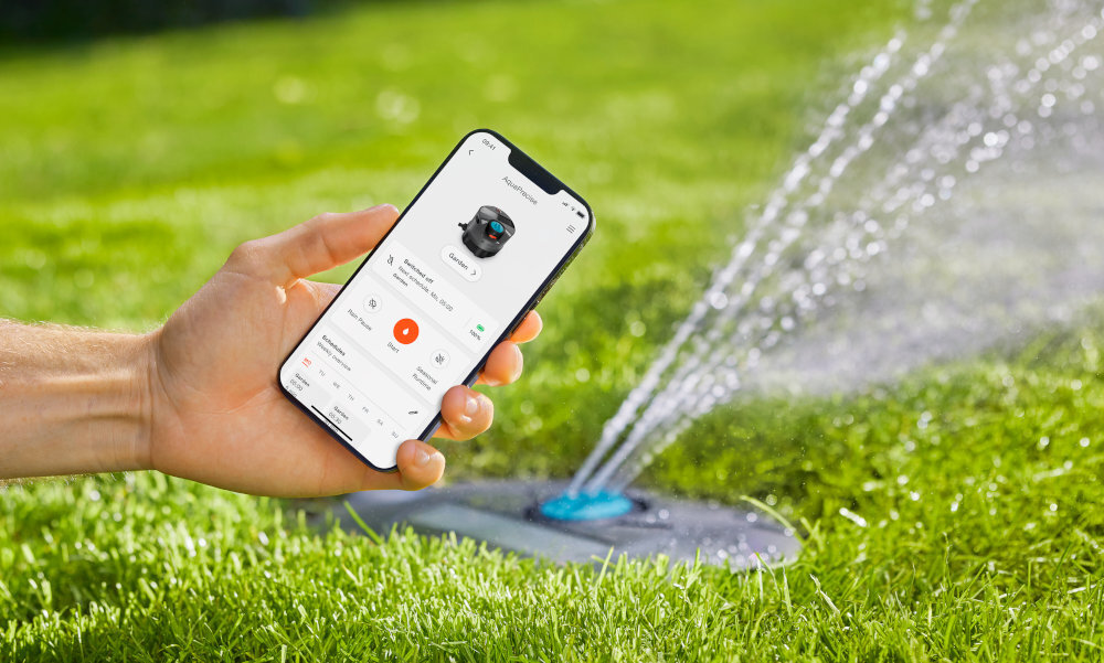 Zraszacz GARDENA AquaPrecise 16001-20 osoba trzyma telefon w dłoni z włączoną aplikacją w tle trawnik trawa zraszacz podlewa trawę wbudowany sterownik pełna obsługa systemu za pomocą aplikacji GARDENA Bluetooth® można tworzyć harmonogramy pracy dopasowywać czas podlewania monitorować przepływ wody zasięg do 10 metrów