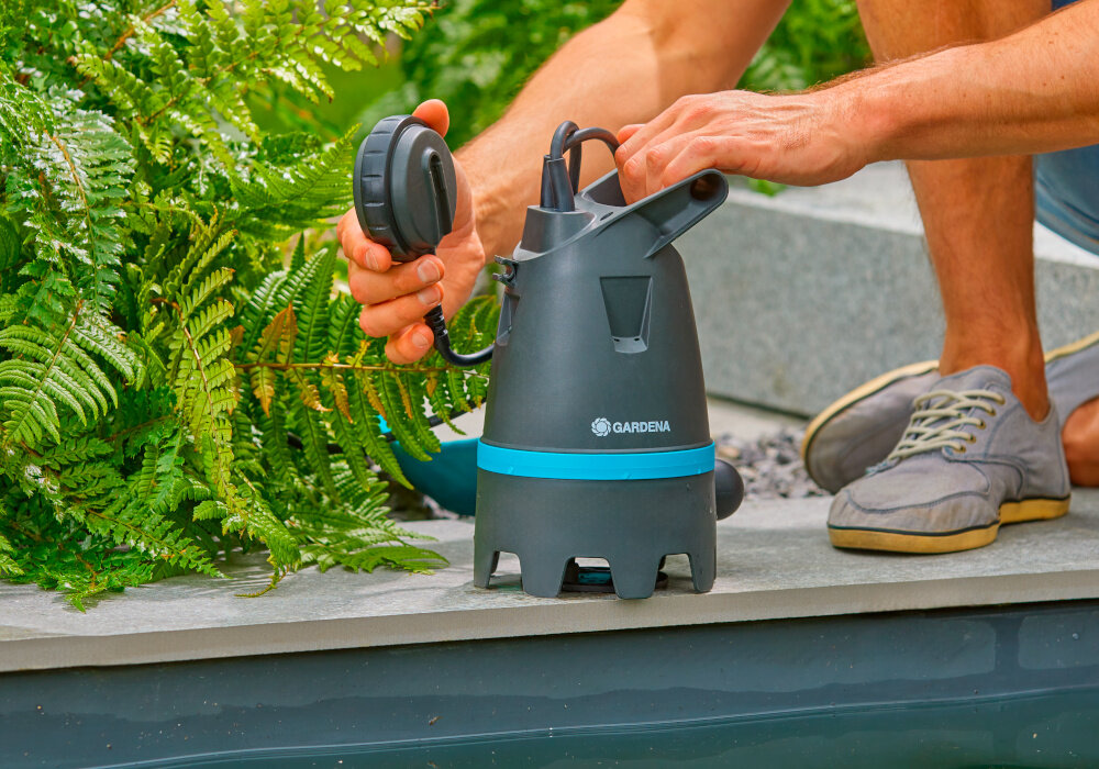 Pompa zanurzeniowa do wody brudnej GARDENA 10500 Basic 09008-47 elektryczna brzeg oczka wodnego pompa na brzegu wokół rośliny zielone osoba trzyma pompę w dłoniach szeroki zakres zastosowań do czystej i brudnej wody cząstki stałe nie przekraczają 25 mm średnicy