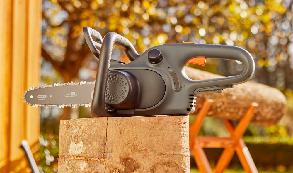 Piła akumulatorowa GARDENA PowerSaw 14790-55 piła leży na drewnianej kłodzie w tle drzewa wygodna praca w ogrodzie precyzyjny łańcuch zaawansowane rozwiązania łatwe cięcie gałęzi przygotowywanie drewna opałowego i pielęgnację drzew wygodę i bezpieczeństwo